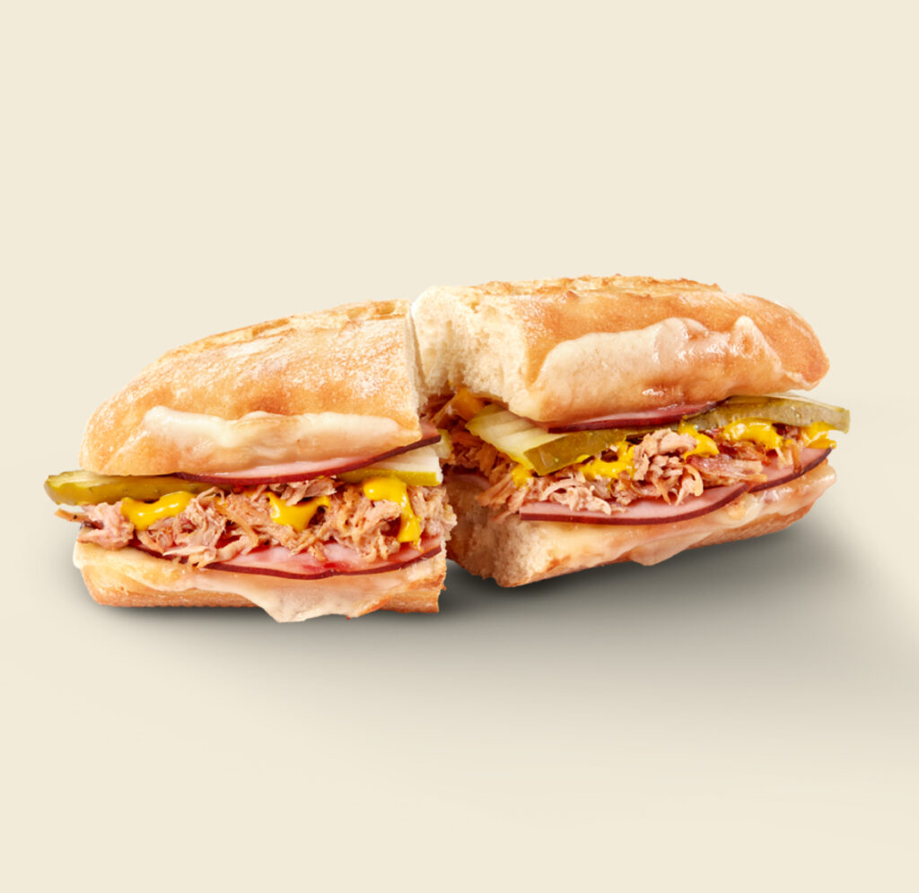 The Cuban Sandwich, featuring slow-roasted pulled pork, ham, Swiss cheese, crunchy pickles, and a drizzle of mustard on freshly baked artisan bread.