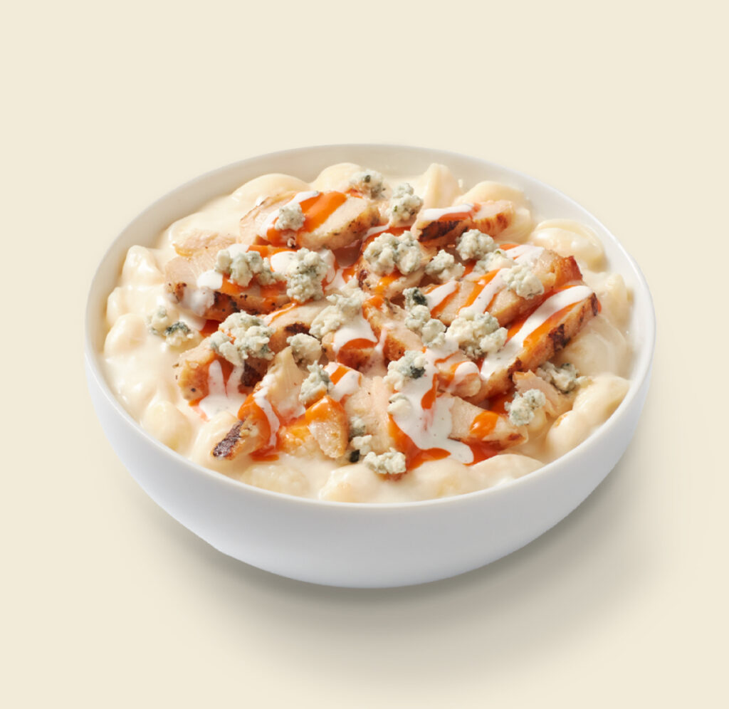 A bowl of creamy mac and cheese topped with grilled chicken, bleu cheese crumbles, buffalo sauce, and ranch dressing on a light background.