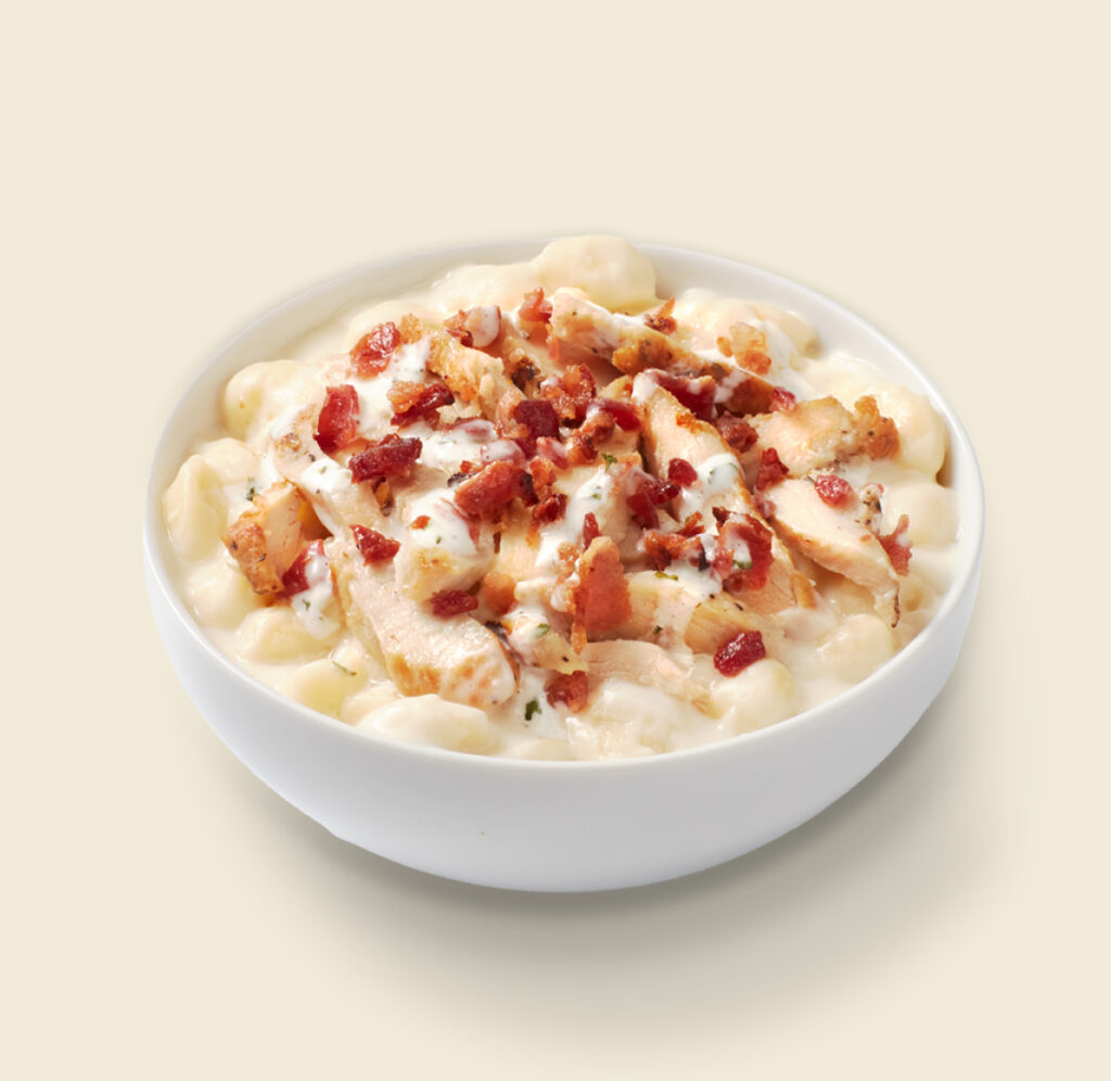 A bowl of creamy mac and cheese topped with grilled chicken, crispy bacon, and ranch dressing on a light background.